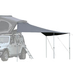 A black rooftop tent with an extended gray tarp attached to it, set up outdoors.