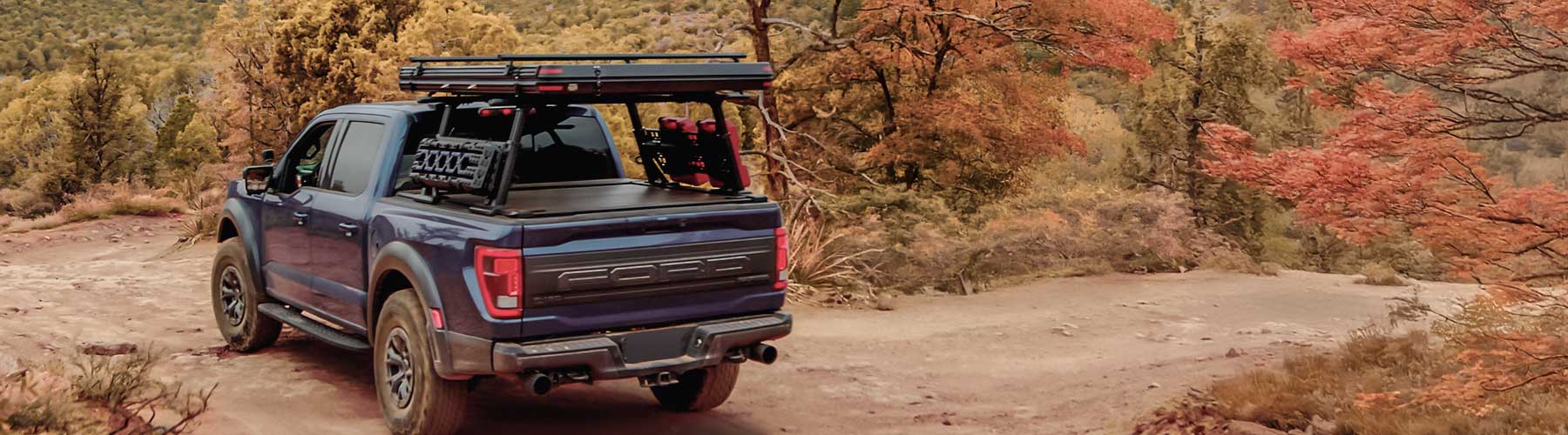 Blue truck with a roof rack on a dirt road surrounded by autumn trees.
