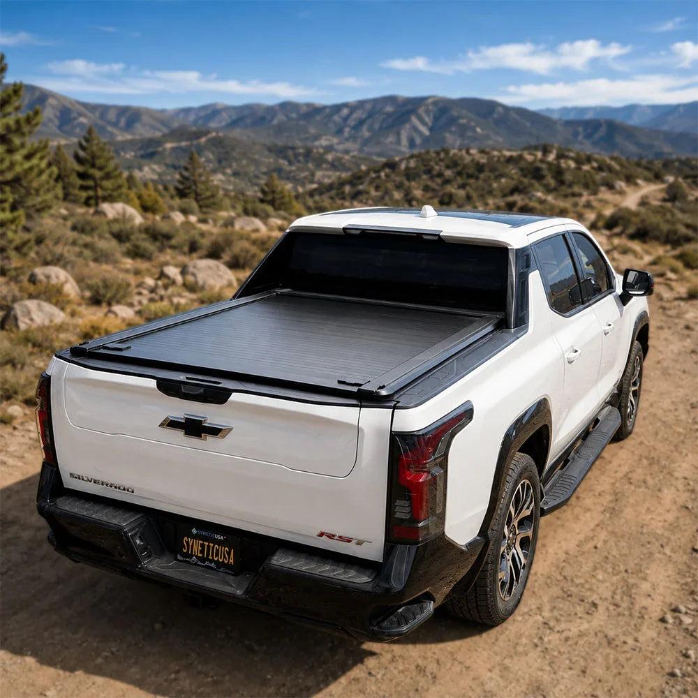 A white Chevrolet Silverado EV truck parked on a dirt road with mountains and trees in the background. The truck bed is covered by a black tonneau-style cover, shown from the rear three-quarter angle.