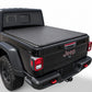 A black soft roll-up tonneau cover installed on the bed of a black Jeep Gladiator pickup truck, shown from a rear three-quarter angle against a light gray studio background.