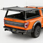 An orange Ford pickup truck shown from the rear three-quarter angle with a black hardtop canopy mounted over the bed and rear window area, photographed in a clean studio setting.