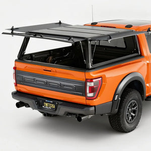 An orange Ford pickup truck shown from the rear three-quarter angle with a black hardtop canopy mounted over the bed and rear window area, photographed in a clean studio setting.