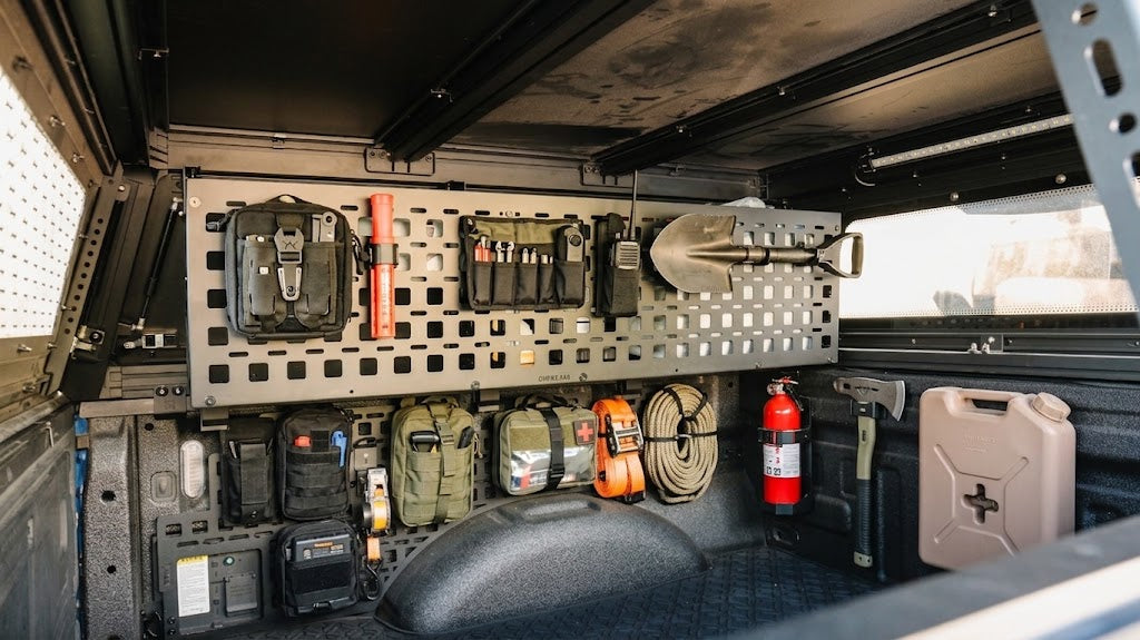A tactical vehicle storage compartment with a metal grid rack holding gear, including black pouches, an orange flashlight, a shovel, a fire extinguisher, coiled rope, and a beige fuel canister.