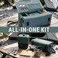 An outdoor camping cookware set displayed on a wooden table with a tent in the background. The set includes a large sage green carrying case, a matching multi-level pot, metal utensils, a cutting board, and cookware.