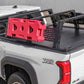 A white pickup truck bed with a black tonneau cover and black metal rack system, carrying a bright red molded plastic cargo bag. The truck shows a visible 'TRD Off Road' badge on the rear quarter panel.