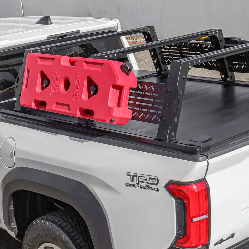 A white pickup truck bed with a black tonneau cover and black metal rack system, carrying a bright red molded plastic cargo bag. The truck shows a visible 'TRD Off Road' badge on the rear quarter panel.