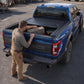 A man in work clothes stands beside a blue pickup truck in an outdoor lot, reaching into the bed where a black tonneau-style bed cover is installed. The truck tailgate shows a large '10' badge.