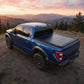A blue Ford pickup truck parked outdoors on a dirt trail with mountains and trees in the background at sunset. The truck bed is covered by a black tonneau-style cover.