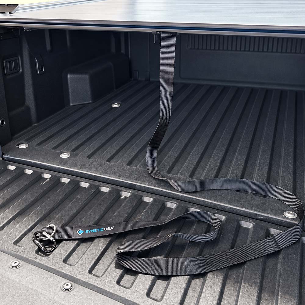Black truck bed liner strap with a blue and white SYNTHETIC USA logo, laid out on the textured black bed liner of a pickup truck.