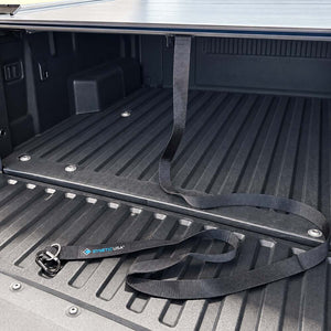 Black truck bed liner strap with a blue and white SYNTHETIC USA logo, laid out on the textured black bed liner of a pickup truck.