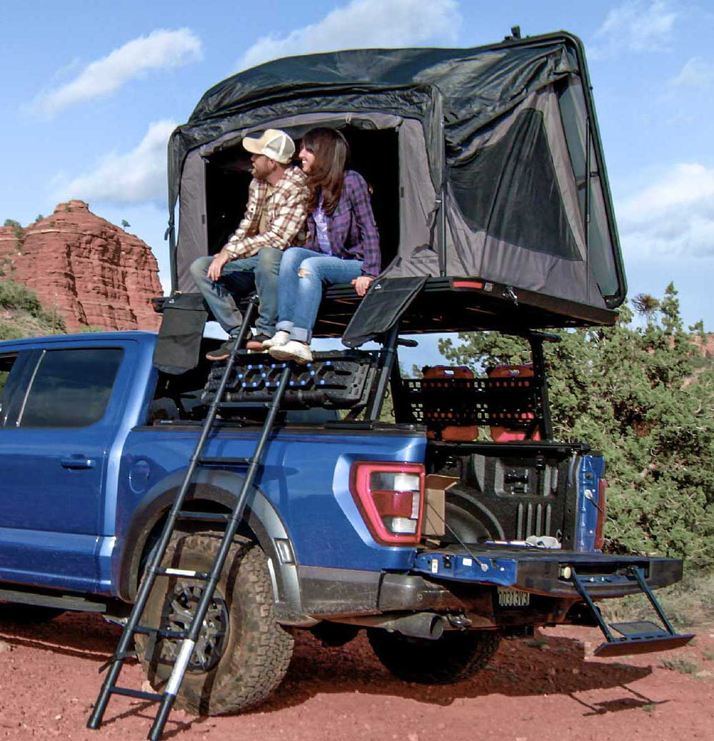 Two people sitting in a rooftop tent on a blue truck with a scenic background