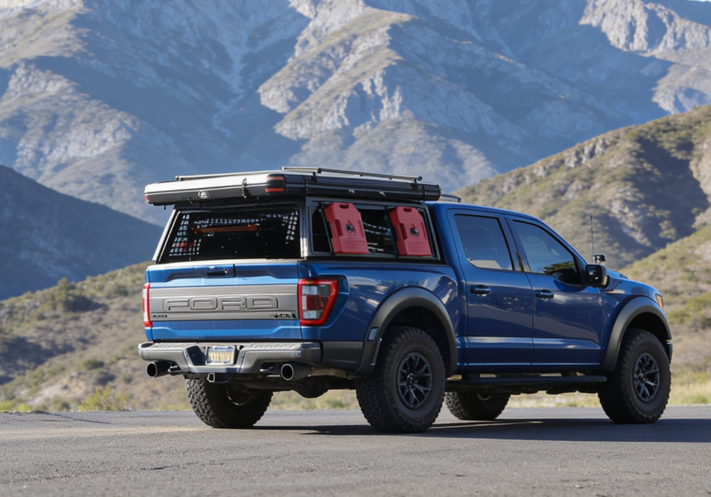 A blue Ford pickup truck with a black roof rack and red side cargo carriers, parked on a paved road with mountains in the background.