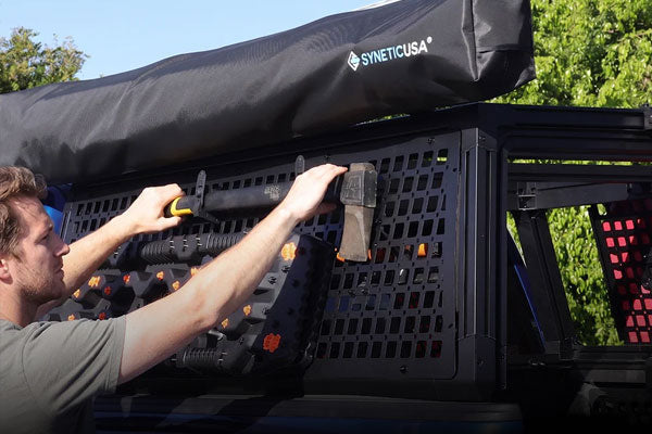 A black Syneticusa truck bed rack system mounted on the bed of a blue truck, holding a rifle and an axe. A person's arm is visible on the left pointing toward the rack.