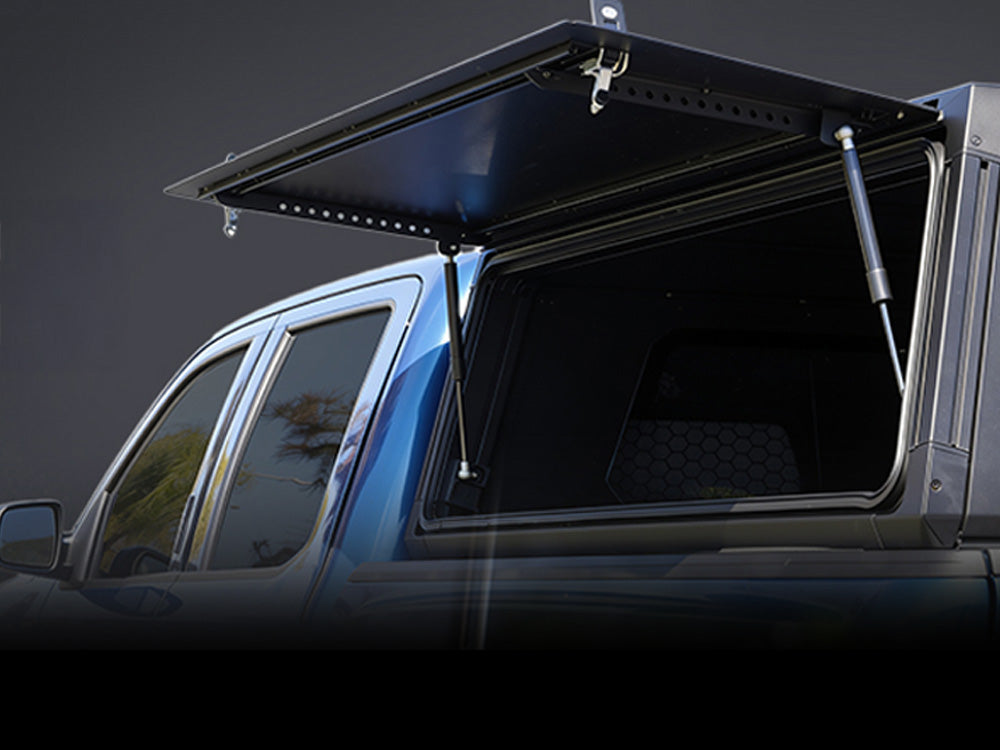 A black hardtop truck bed cover with hydraulic support arms open, mounted on the bed of a blue pickup truck against a dark studio background.