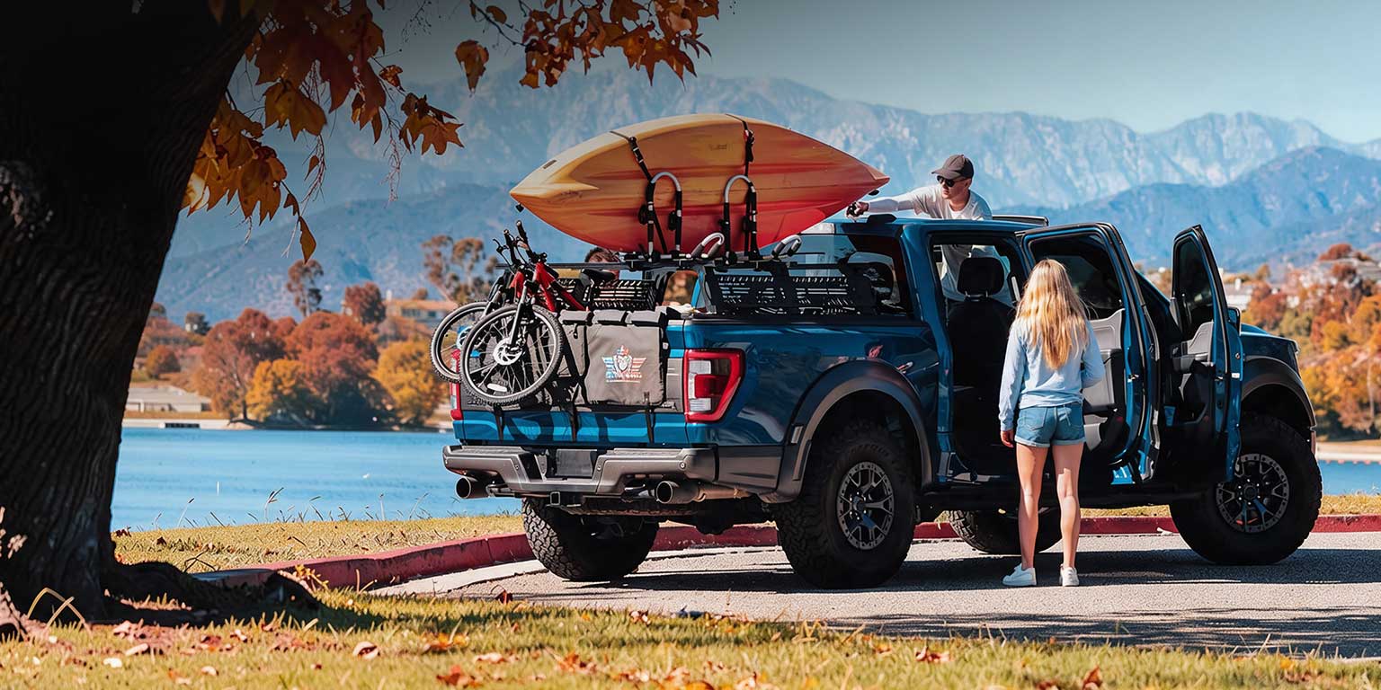 A blue pickup truck parked outdoors by a lake with mountains in the background. A person is loading a surfboard onto the truck bed while another stands by the open driver's side door.