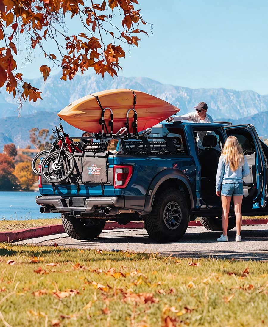 A blue pickup truck parked outdoors with a person loading/unloading from the open rear door and another person at the truck bed. The truck is loaded with a large orange surfboard on top, a bicycle in the bed, and gear racks.