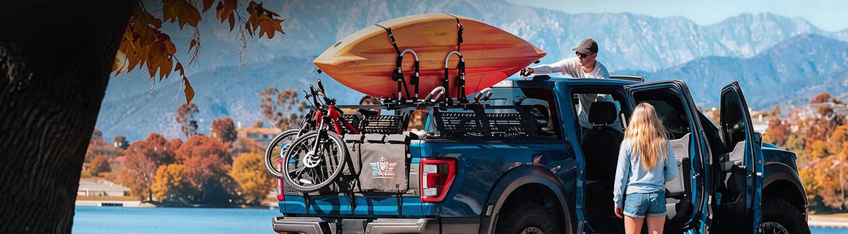 A blue pickup truck parked outdoors by a lake with mountains in the background. The truck bed is loaded with a surfboard, bicycles, and gear, and two people are standing near the open rear doors.