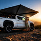 A man stands beside a white pickup truck parked on a rocky mountain trail at sunset, using a large black camping canopy attached to the truck bed. The background shows a glowing mountain landscape under a blue sky.