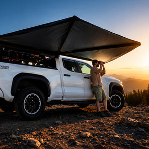 A man stands beside a white pickup truck parked on a rocky mountain trail at sunset, using a large black camping canopy attached to the truck bed. The background shows a glowing mountain landscape under a blue sky.