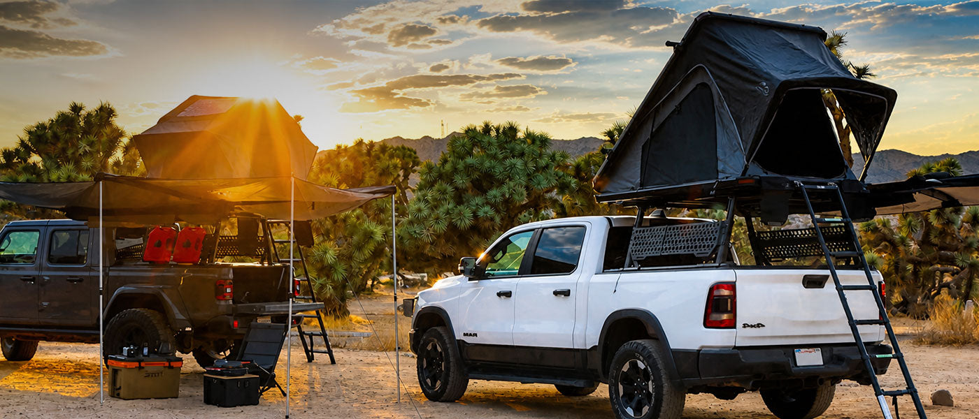A white Ram pickup truck with a rooftop tent and ladder setup in a desert campsite at sunset, alongside another vehicle with an awning and camping gear.