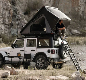 Person sitting on a ladder accessing a rooftop tent on a Jeep in a natural setting.