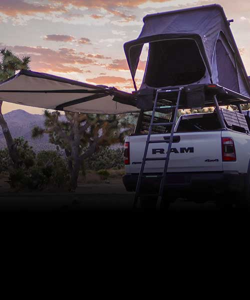 White RAM truck with a rooftop tent and extended awning in a desert landscape at sunset.