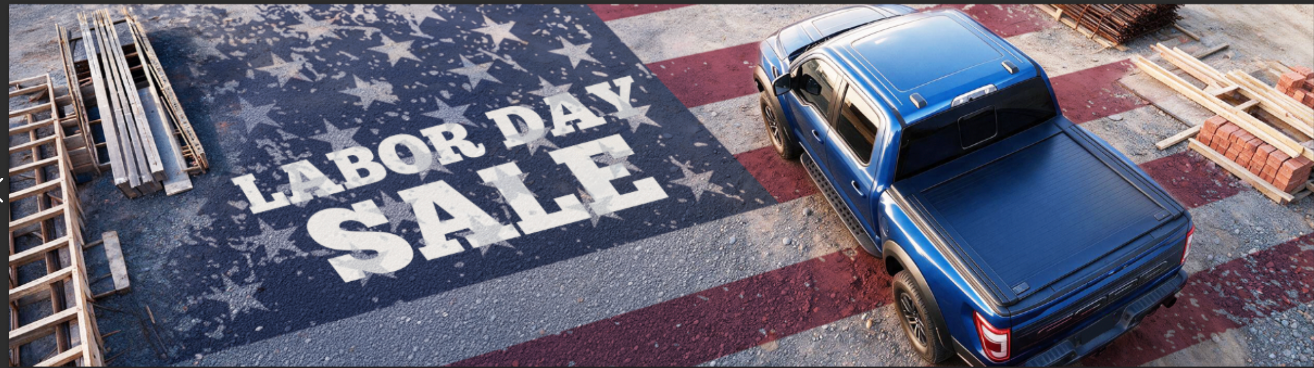 A blue pickup truck parked on a construction site with a large 'LABOR DAY SALE' sign painted on the ground, surrounded by wood stacks and materials.