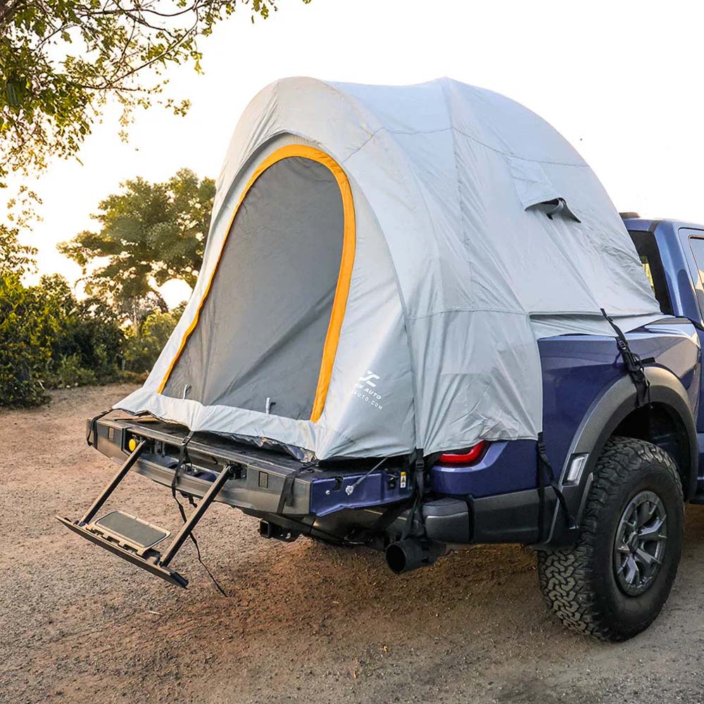 A light gray rooftop tent with a yellow-trimmed front window is mounted on the bed of a dark blue pickup truck, parked outdoors on a dirt road with trees in the background.