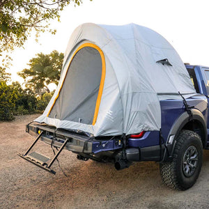 A light gray rooftop tent with a yellow-trimmed front window is mounted on the bed of a dark blue pickup truck, parked outdoors on a dirt road with trees in the background.