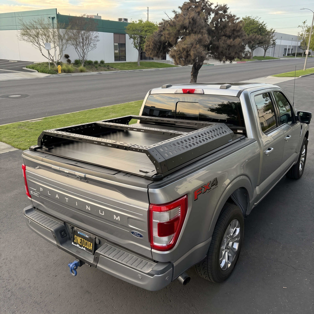 A silver Ford F-150 Platinum pickup truck parked outdoors in a parking lot, shown from the rear three-quarter angle. The truck bed is fitted with a black metal tonneau-style cargo bed cover and a black diamond-plate style panel on top.
