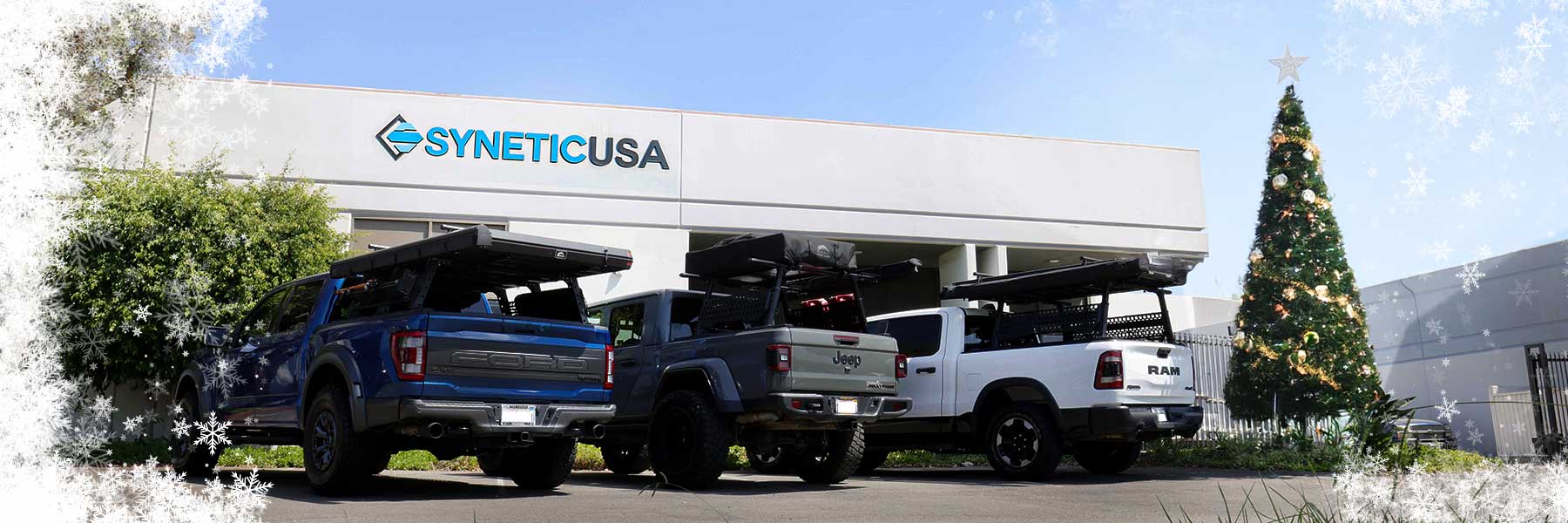 Two pickup trucks parked in front of a building with 'SYNETICUSA' branding, Christmas tree in the background.