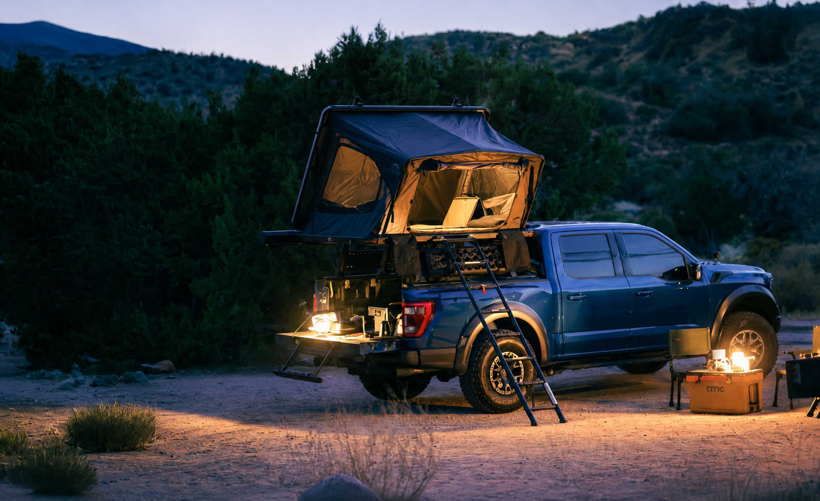 Blue truck with a rooftop tent parked in a desert landscape at dusk.