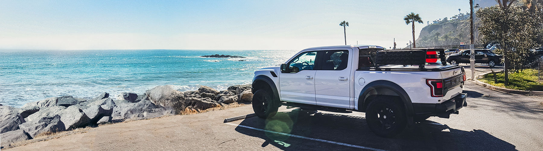 White f150 raptor truck parked on a road near a coastal area with ocean and rocks in the background.