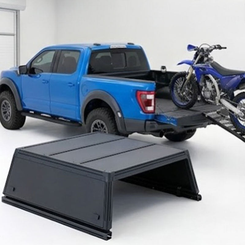 A black metal truck bed ramp and cover accessory shown in the foreground, with a blue pickup truck and a blue motorcycle in the background on a white studio floor.