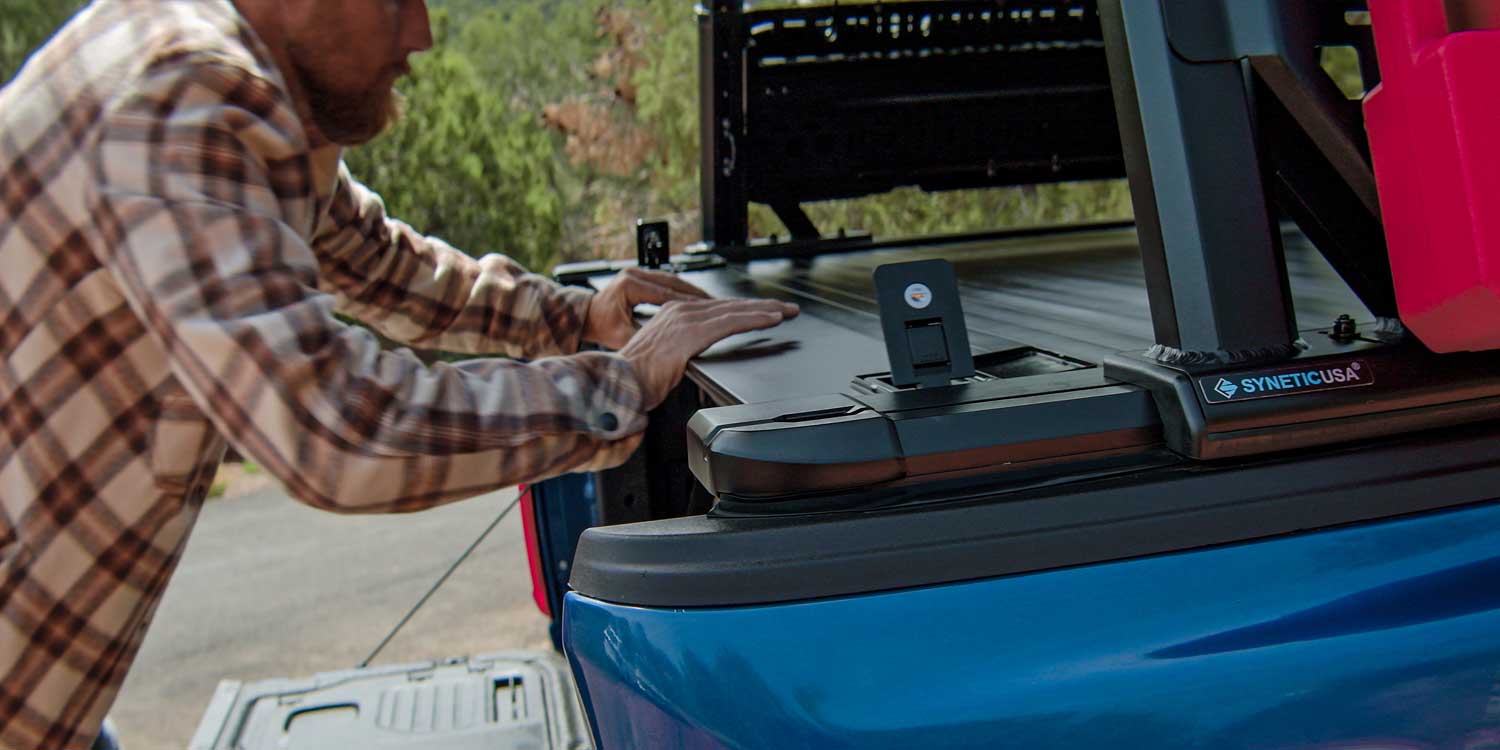 Person adjusting a tonneau cover on the back of a blue truck with a scenic background