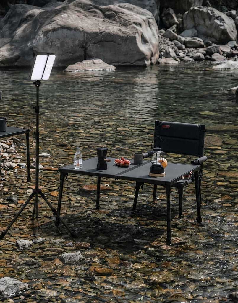 Table setup with a chair by a body of water, including a light stand and various items on the table.
