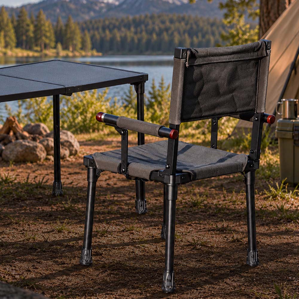 A green camp chair with fabric seat and back is shown outdoors beside a folding table, with a lake, trees, and mountains in the background.