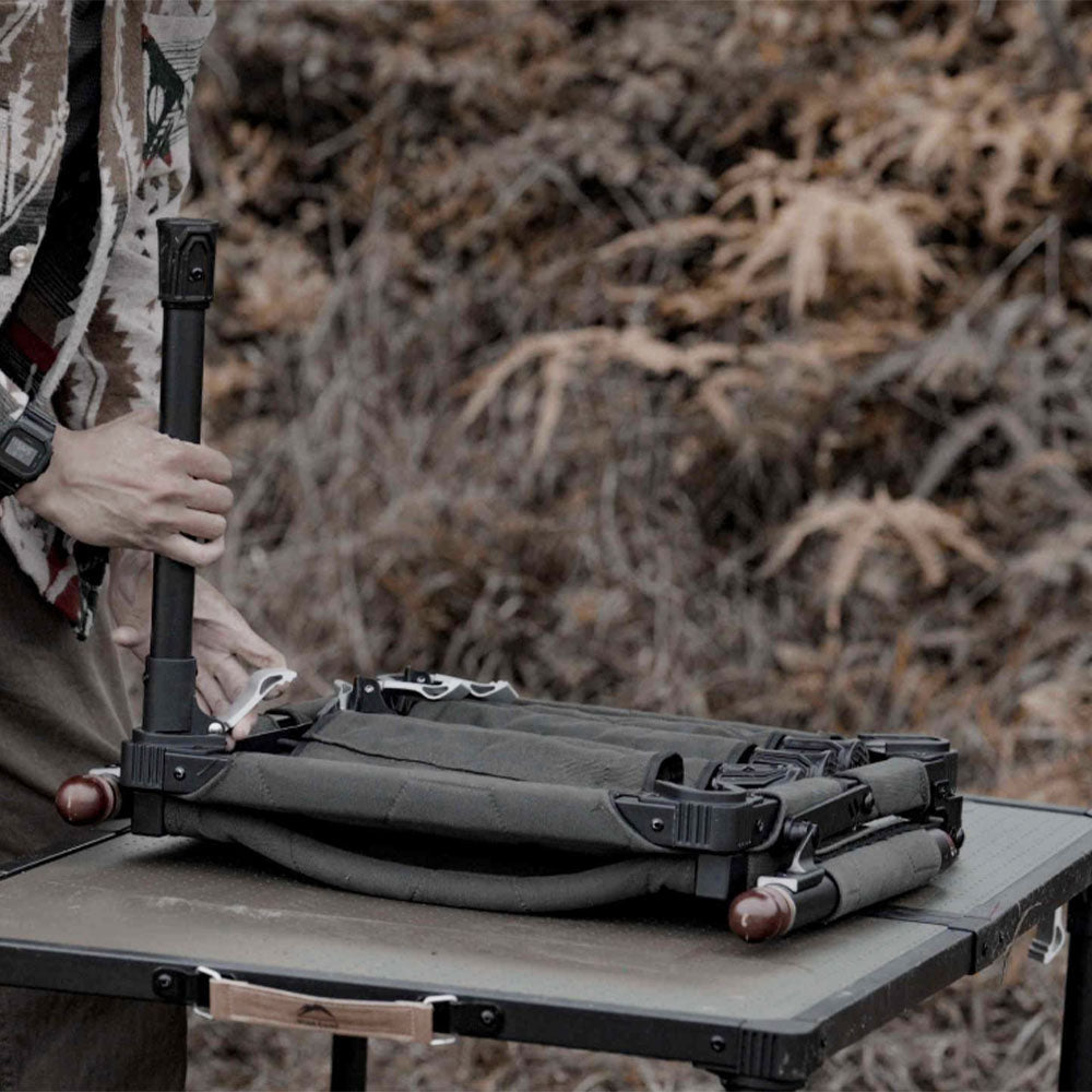 A person outdoors adjusts a green folding camp cot on a small table, with dry brush in the background.