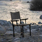 A folding camp chair with a canvas seat and backrest stands on a rocky shore next to a body of water at sunset.