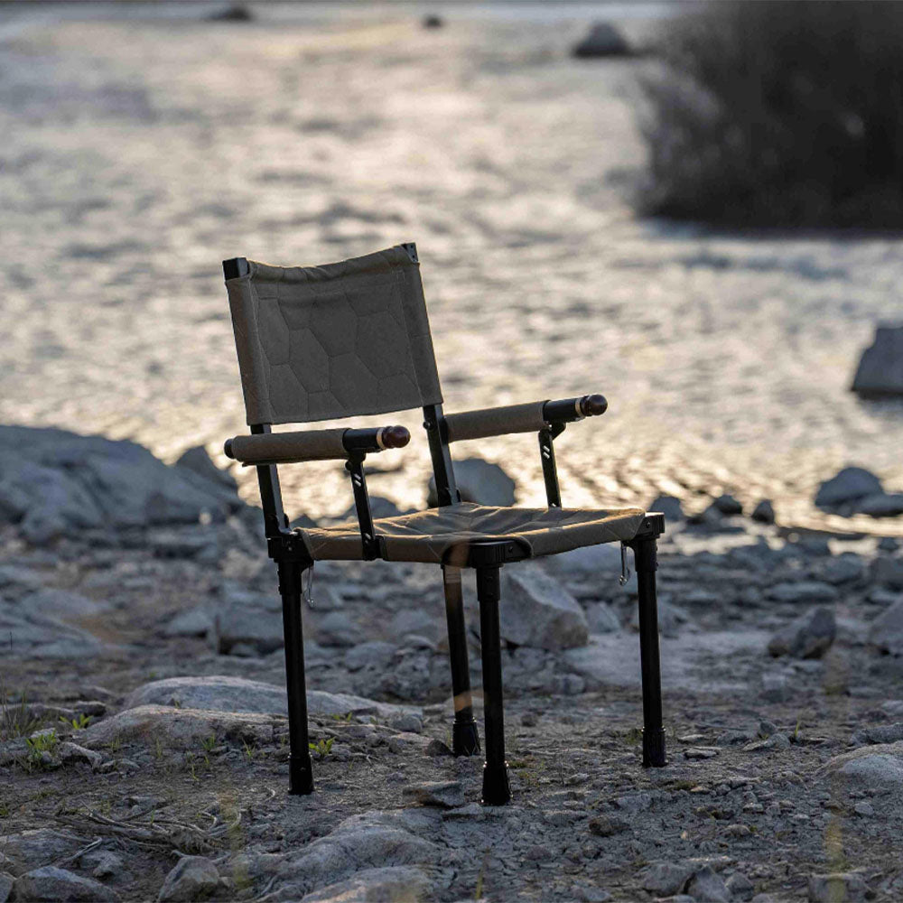 A folding camp chair with a canvas seat and backrest stands on a rocky shore next to a body of water at sunset.