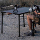 A black folding camp table set outdoors on gravel near a person seated at one end, holding a camera. A lantern sits on the table, and dry brush is visible in the background.