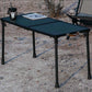 A black folding camp table with a textured top surface, shown outdoors on sandy ground with a lantern and magazine on top.