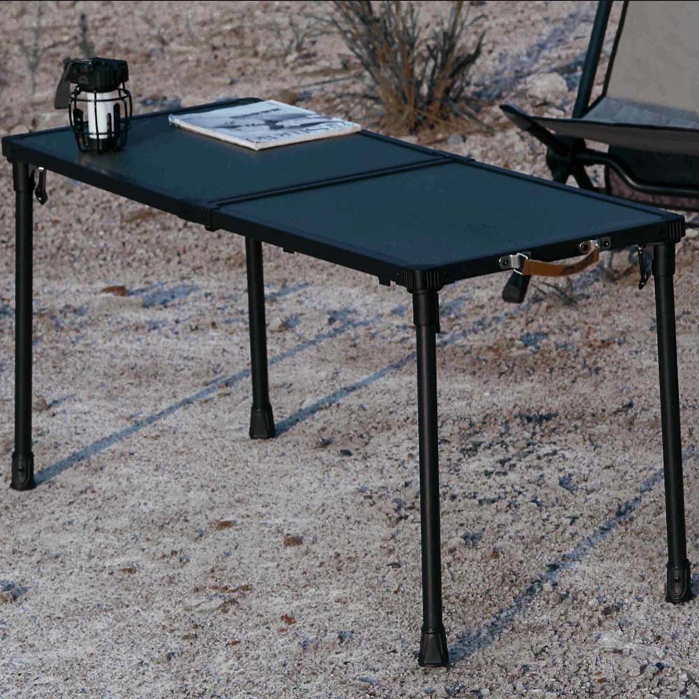 A black folding camp table with a textured top surface, shown outdoors on sandy ground with a lantern and magazine on top.