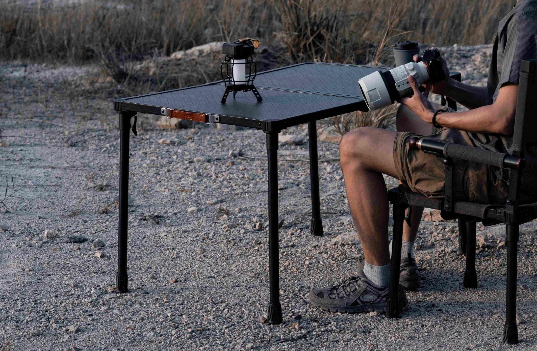 Person sitting at a table outdoors with binoculars and a camera on a tripod.