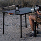 Person sitting at a table outdoors with binoculars and a camera on a tripod.