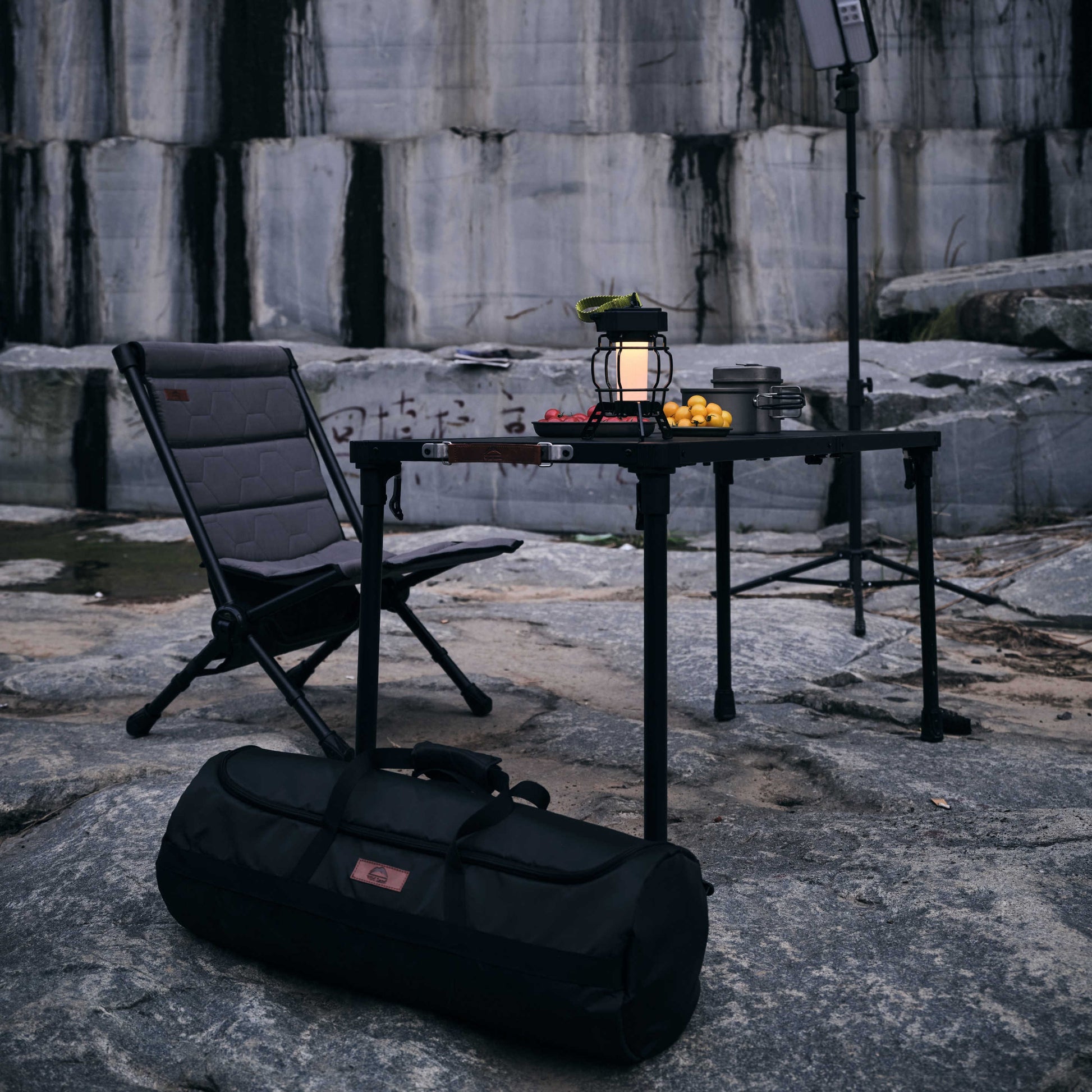 Camping setup with chair, table, lantern, and duffel bag against a rocky background