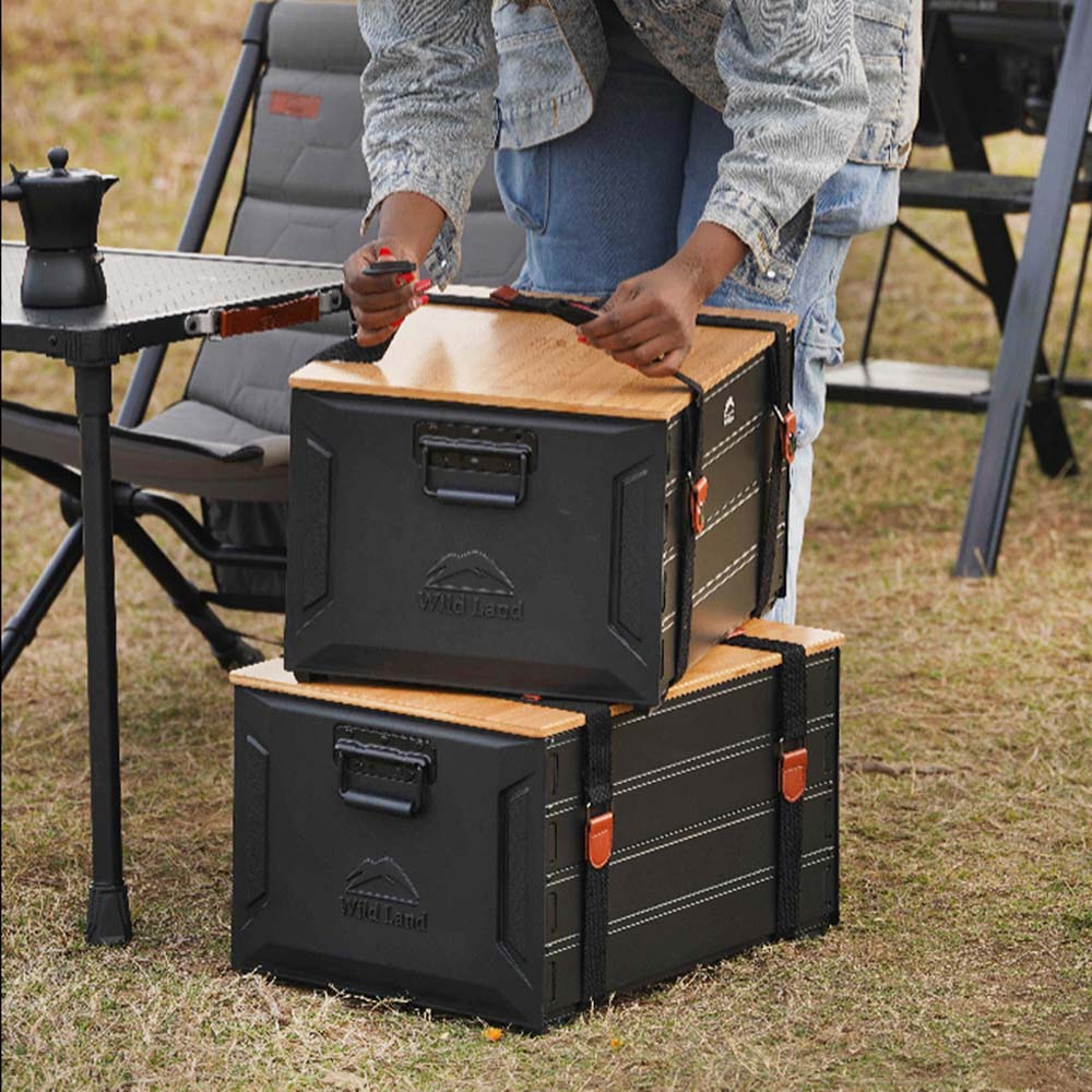 Two outdoor storage boxes with light wood tops are stacked on grass near camping chairs and a table. A person is visible from the waist down adjusting the top box.