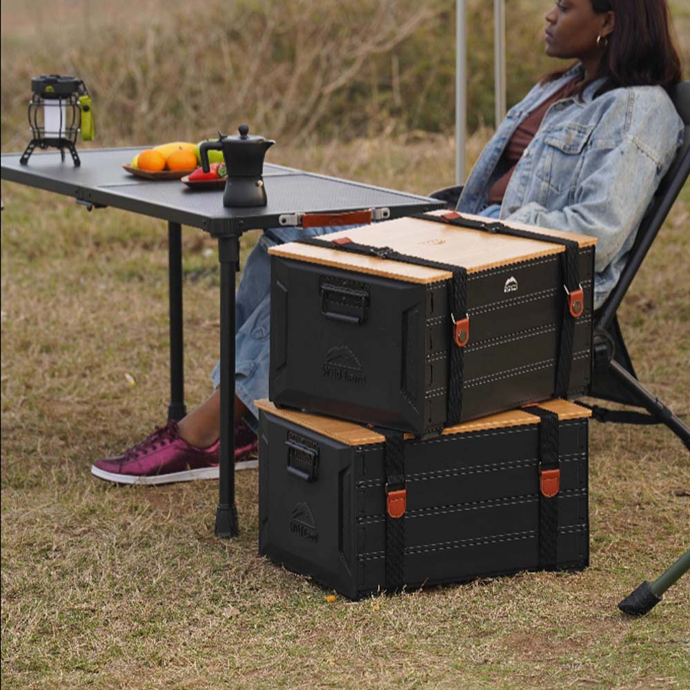 Two outdoor storage boxes with light wood tops are stacked on grass near camping chairs and a table.  